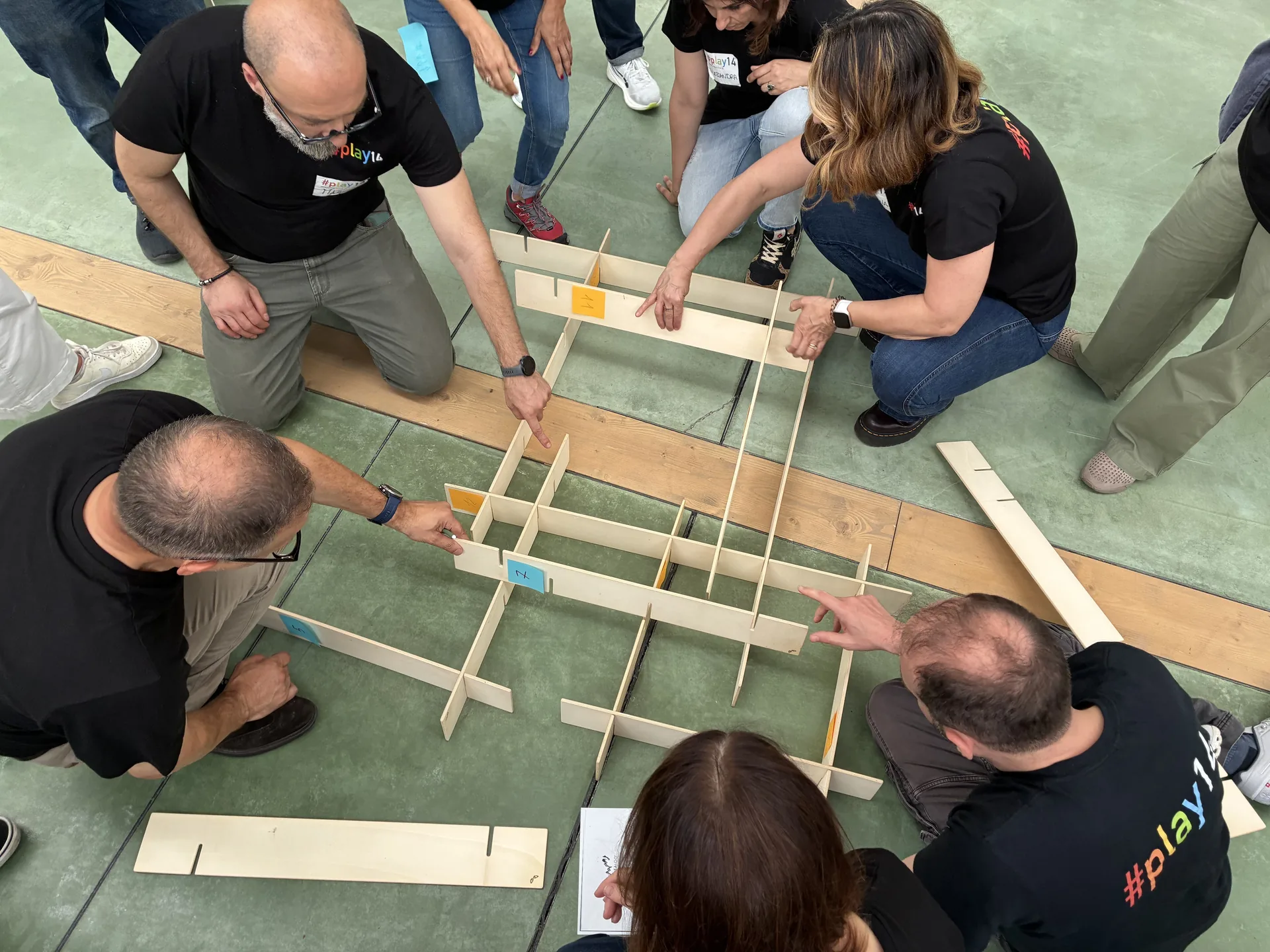Un gruppo di partecipanti, inginocchiati a terra, costruisce insieme una struttura a incastro di pannelli di legno durante un’attività di formazione esperienziale.